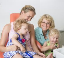 Behandlungszimmer 1, Mutter mit 2 Töchtern im Kleinkindalter, ZÄ Frau Dr. Alexa Frey - Milczewsky in Oberursel – Hochtaunus,größeres Mädchen spielt mit Luft - Wasserspritze am Speibecken, Mutter und kleine Tochter schauen zu.